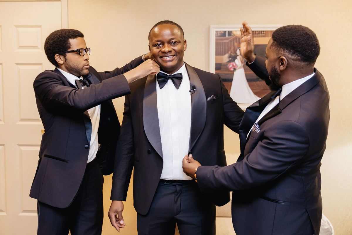 Two of the crew fixing the groom's jacket while he grins